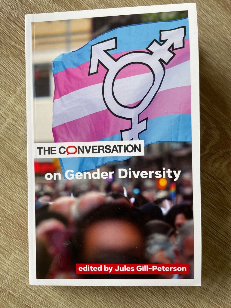 Photograph of a book, The Conversation on Gender Diversity. The cover features a blurred crowd of people and a large trans flag with the trans symbol on top of blue, pink, and white stripes.