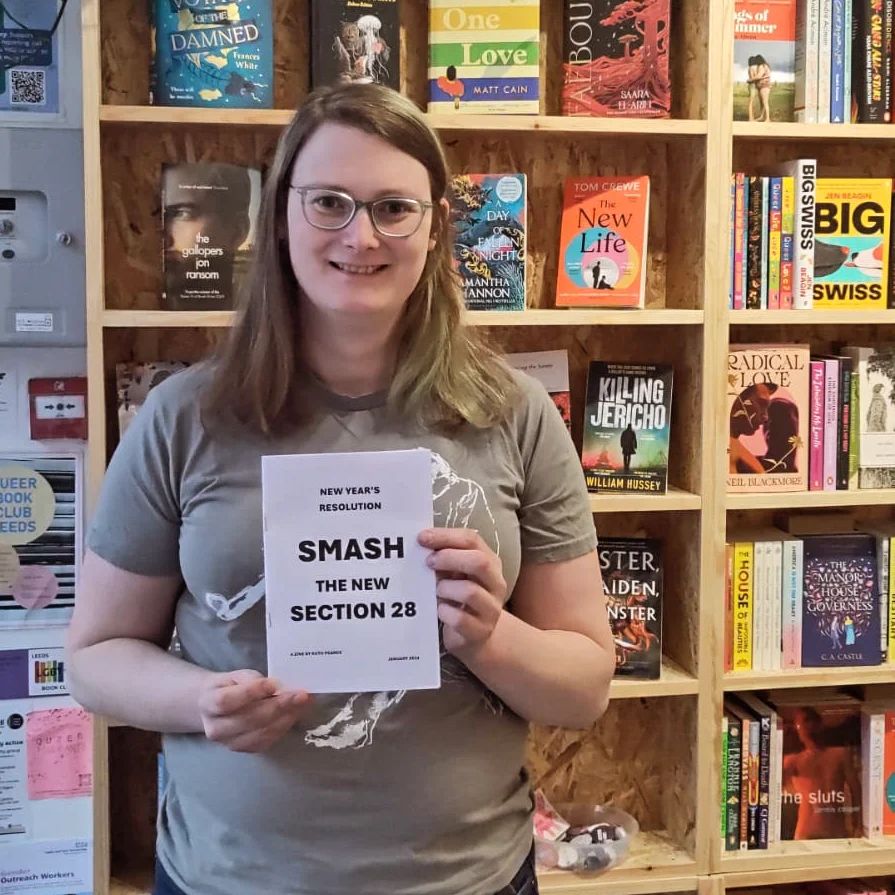 Photograph of a women holding a zine titled Smash The New Section 28. The woman is white and has shoulder-length brown hair, and is wearing glasses and a grey t-shirt. She stands in front of a large book shelf.
